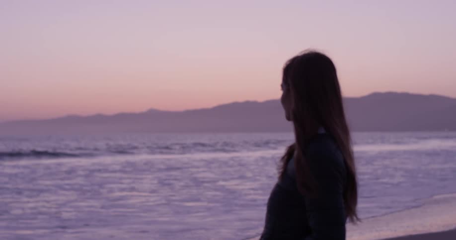 A beautiful young woman stands on the beach in front of a beautiful purple sunset as waves rush in on to the shore with mountains too