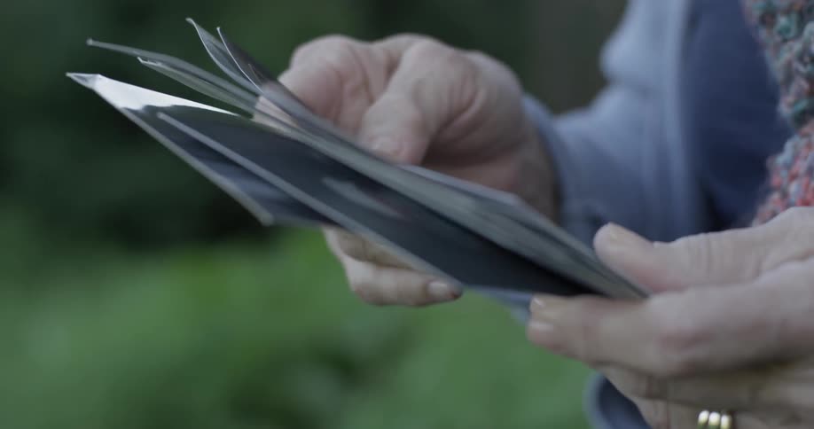 Elderly person sifts through papers perhaps photos with a wedding ring sitting on a park bench on a dim light day