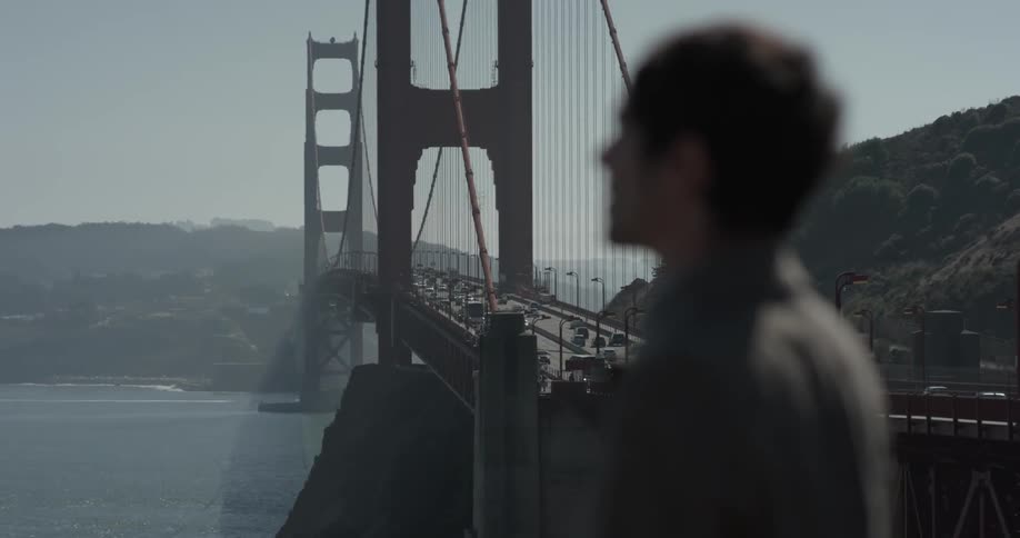 A man looks out over the ocean while standing near a large suspended bridge