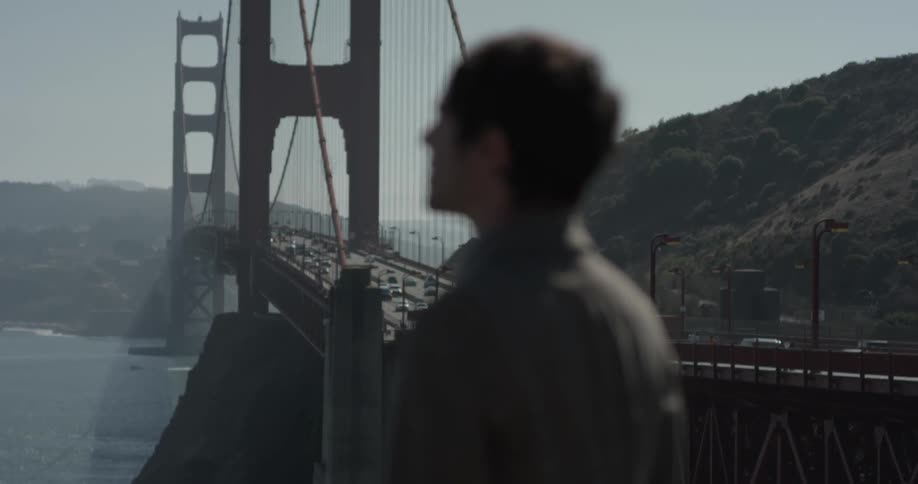 A man stares off into the distance of a red bridge between two large hills on a bright sunny day