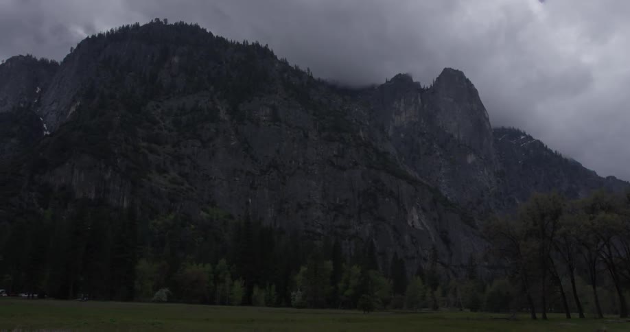 A mountain range is covered in clouds or fog and is adorned with many tall trees and grass fields at the bottom