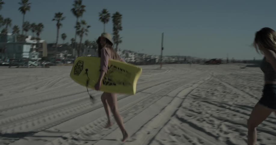 Woman with paddle board runs with another woman on the beach