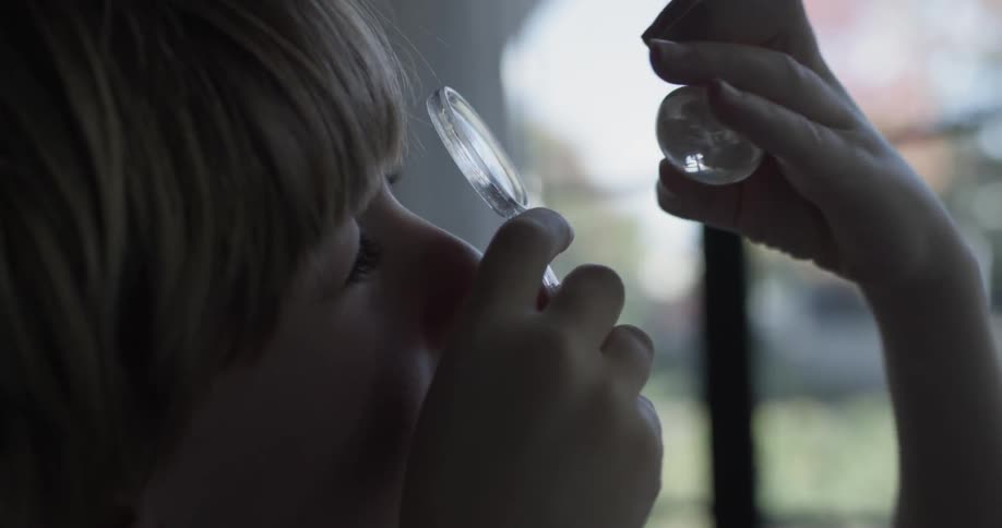 A child inspects a glass oval shaped object with his magnifying glass on a sunny day indoors