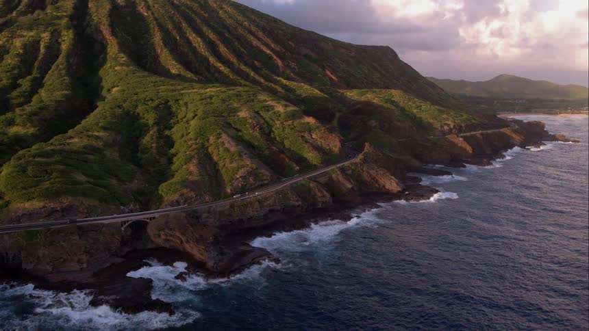Cars drive beside the ocean while overlooked by a tall green hill