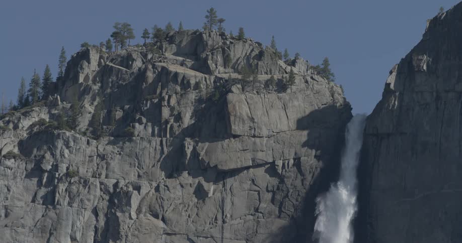 Water flows off of the cliff of a mountain with trees on top and blue skies above