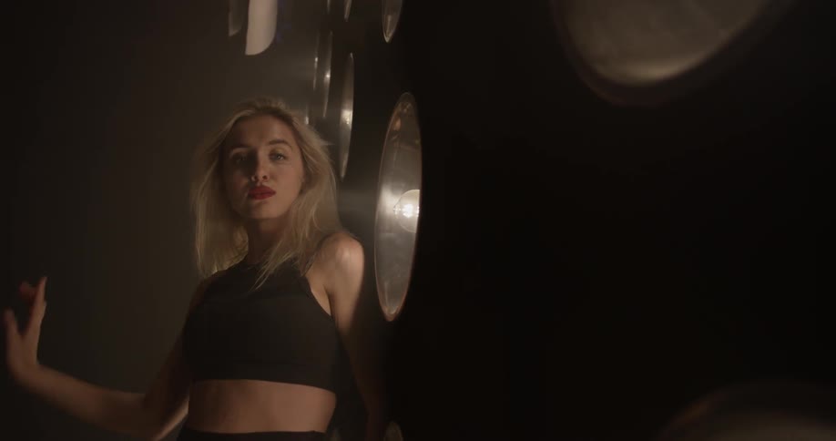 A blond sexy women is seen dancing intimately with light fixtures in the wall in an otherwise dimly lit room