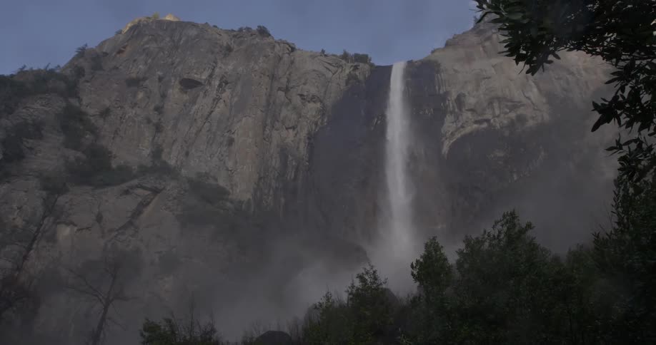 A waterfall has water flowing over the side of a mountain into the rocks below creating mist fog and humidity for the plants and trees below