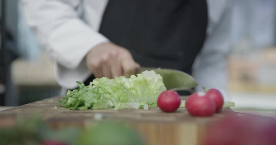 A person cuts lettuce up on the kitchen counter wearing a white shirt and a black apron