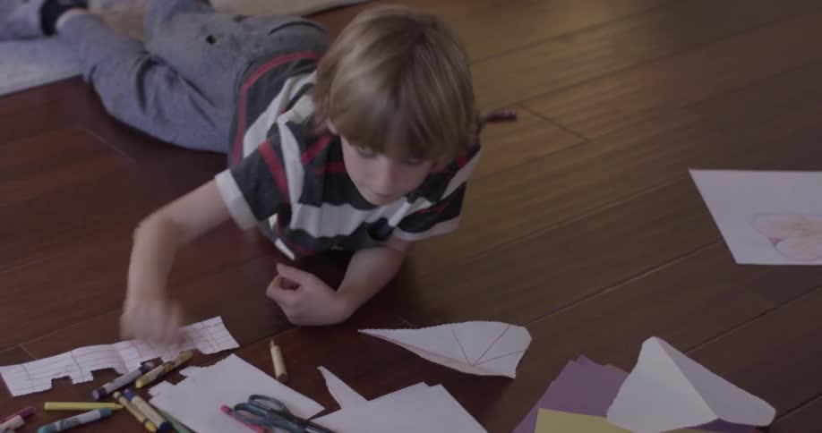 A boy is laying on the floor with many crayons and paper, he is drawing on paper after selecting a crayon