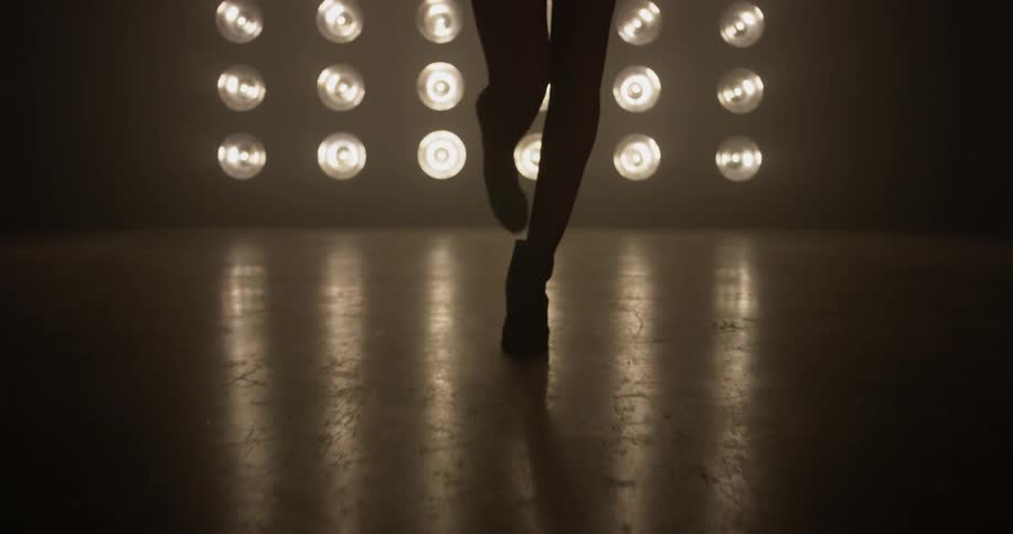 A woman is seen walking towards the camera in a sexy strut wearing boots with lots of lights behind her in an otherwise dark room