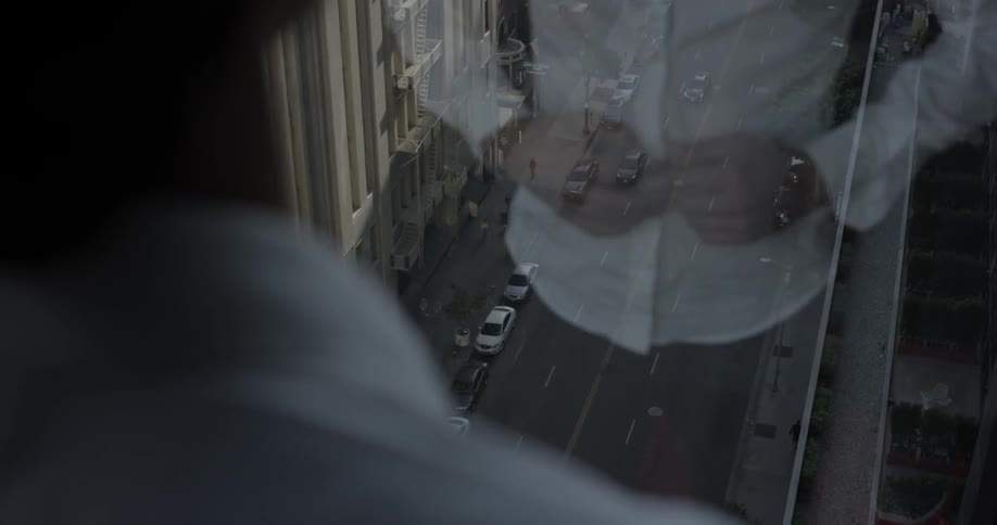 A man stands in front of a large window overlooking a city and buttons his white shirt