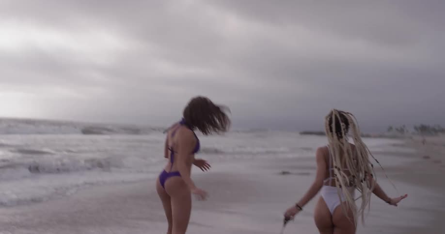 Two women wearing bikinis run and play or have fun pulling a surfboard along down the beach as waves come in on a cloudy day