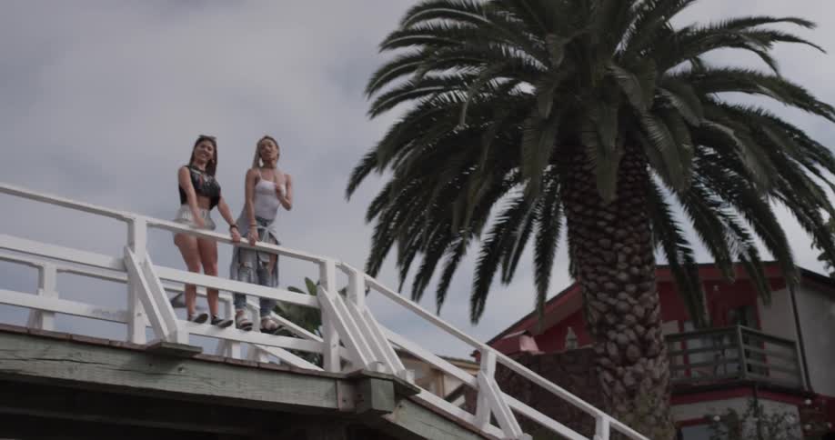 Two women look excited and stand on the railing overlooking a beach front with palm trees on a bright sunny cloudy day