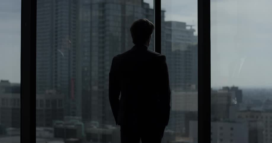 A man stands in the corner of an office room wearing a suit and looking out at the city through tall windows