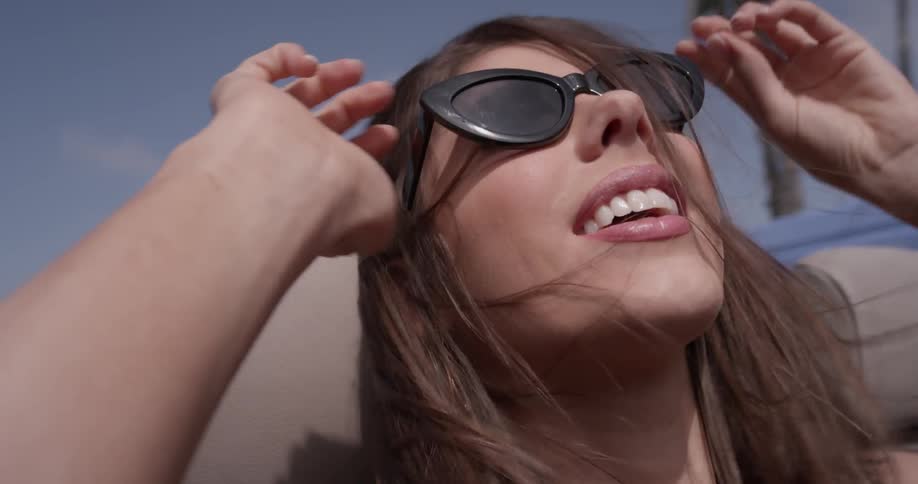 A woman with hair blowing in the wind of a topless car is putting on sunglasses and pursing her lips
