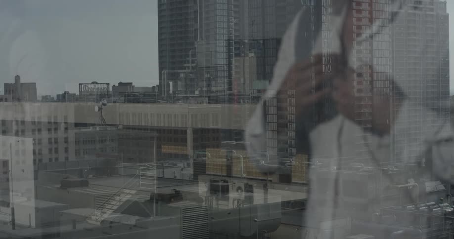 A man buttons up a white shirt as he looks out a window at a city