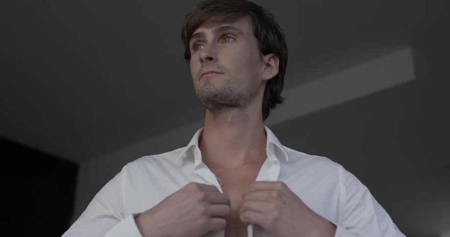 A man puts on a white shirt to button up and stands there staring off into the distance