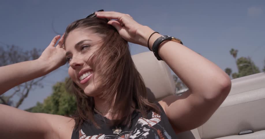 Two women dance in the day with sunglasses on and off adjusting glasses and taking them off with wind pouring in from the open air car