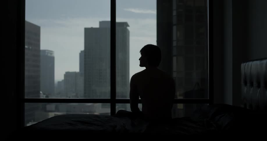 A man sits on his bed overlooking the city outside of the window in a dim lit room