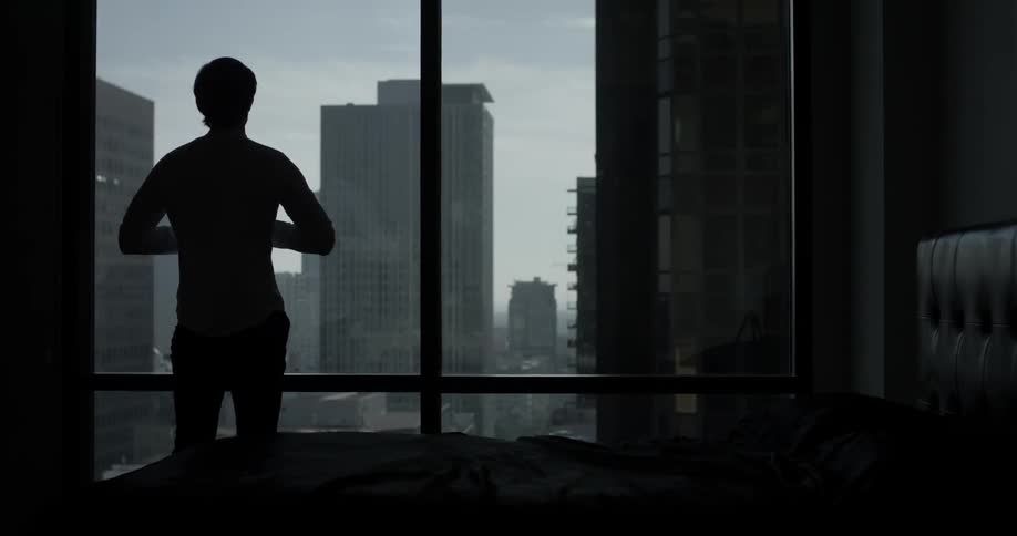 Man gets up out of bed and looks out the tall large windows at the cityscape beyond