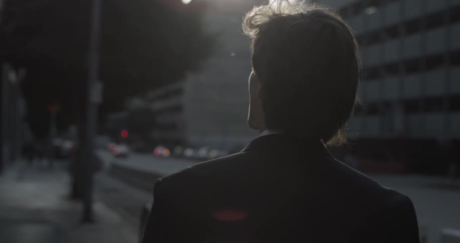 A man walks in slow motion on the sidewalk of a busy city with tall buildings all around him sometime in the late afternoon