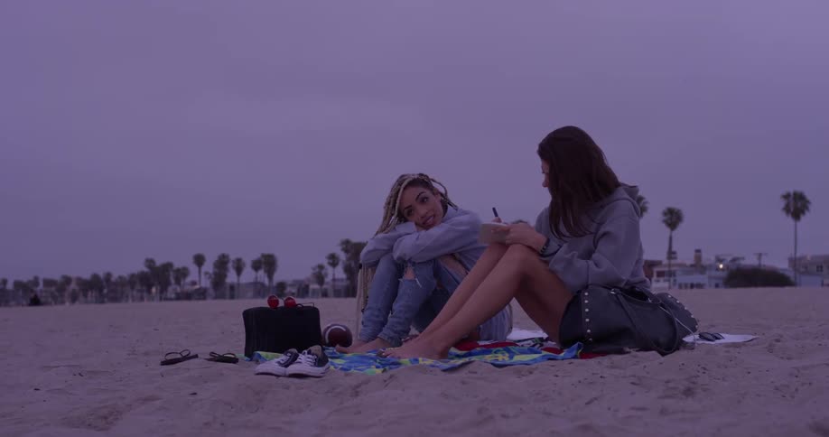 Two women sit at the beach waterfront after the sun has set on a cloudy day with their shoes off and a car passes by