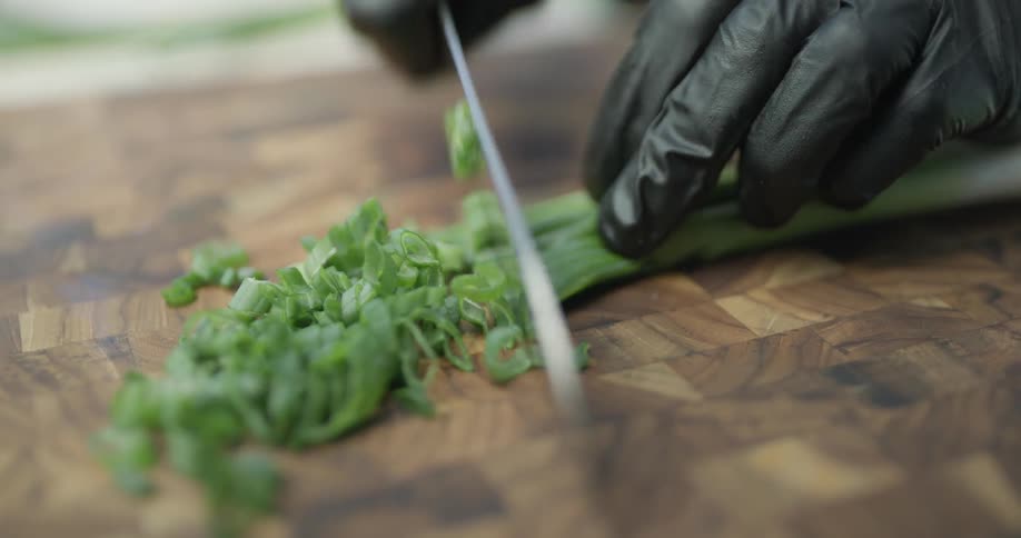 A person wearing black gloves chops up chives on a wooden cutting board with a sharp knife