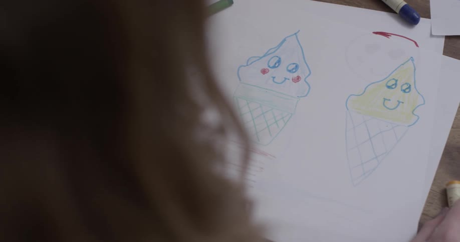 A young girl draws three ice cream cones with faces on a piece of paper