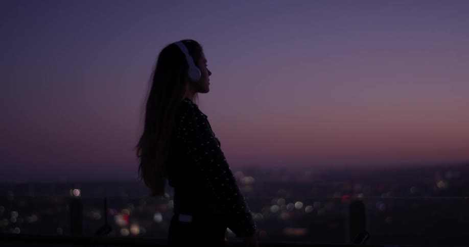 Woman puts headphones on outside at night with a city below
