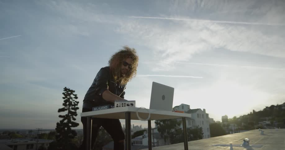 A man hovers over a table with music equipment and a laptop mixing and turning nob's to adjust the music he is playing