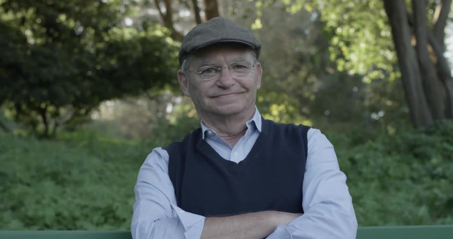 Older man wearing hat sits on bench and looks around as he smiles