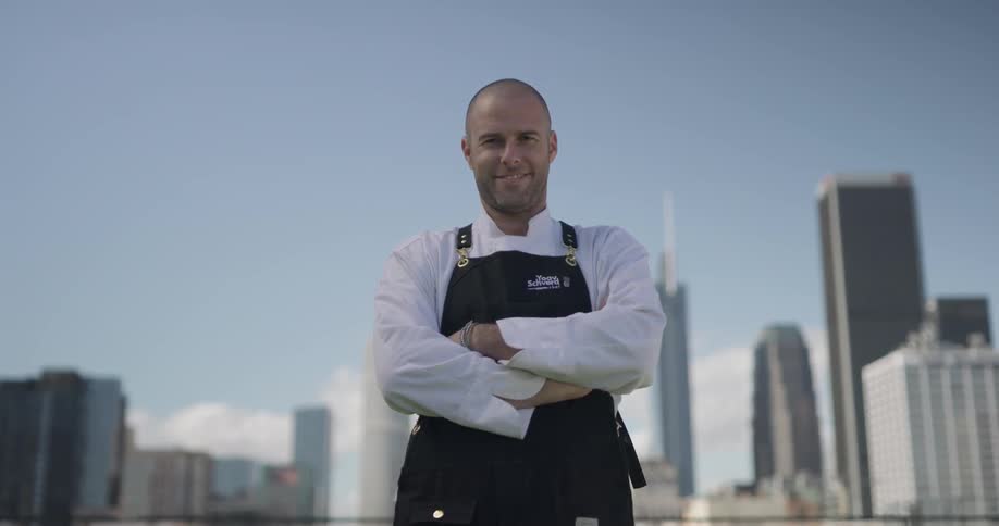 A chef stands on top of a building with a black apron and stares at the camera
