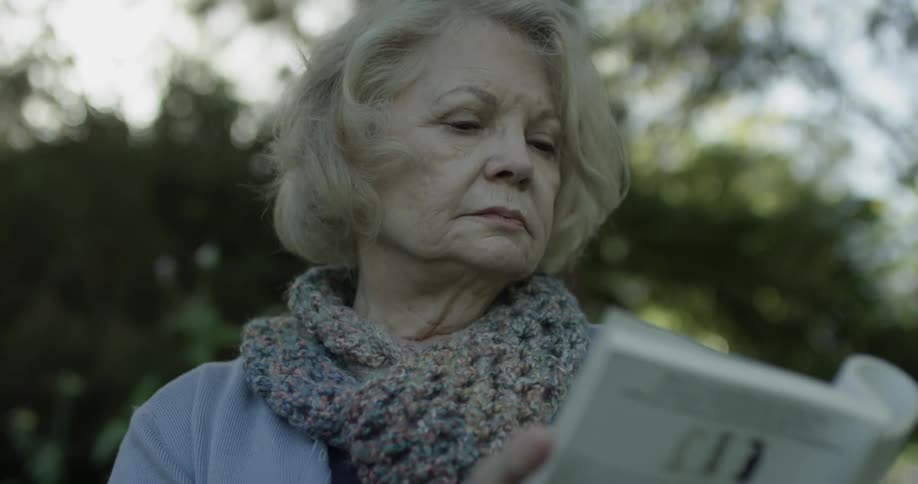 Woman is sitting in the shade of a park on a bench and reading a book to herself