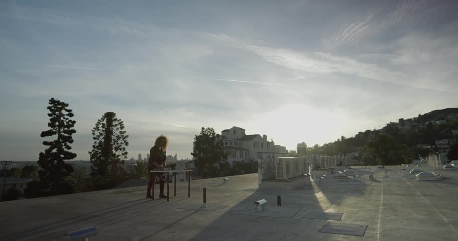 Man stands in front of a laptop and music equipment on a table on top of a roof on a sunny day