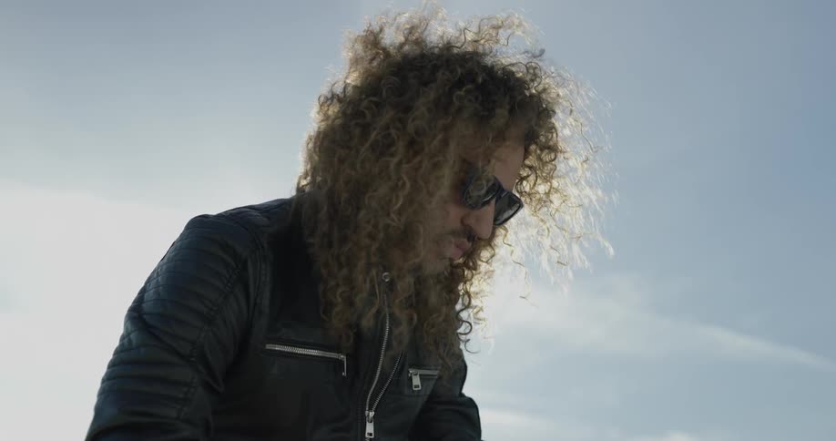 A man with long curly hair stands outside manipulating something with his hands and dancing