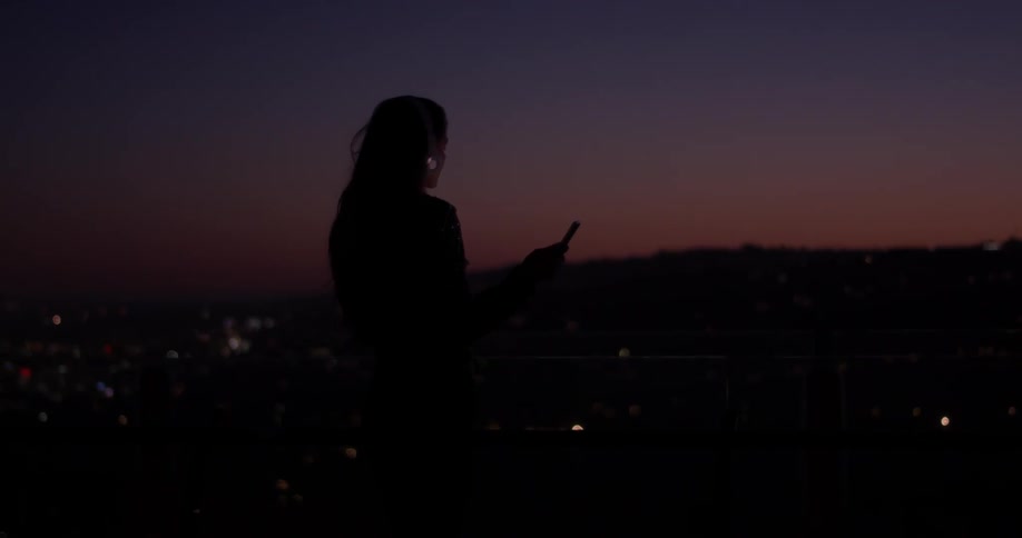 A woman stands with headphones on listening to music and dancing as the sun is setting over the city on top of a hill