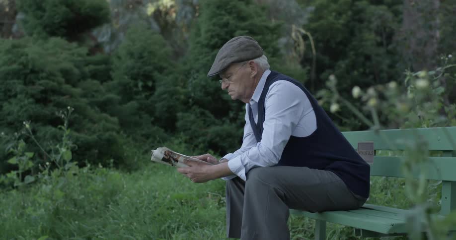 A man sits on a park bench holding a newspaper reading it wearing a hat glasses and a sweater vest
