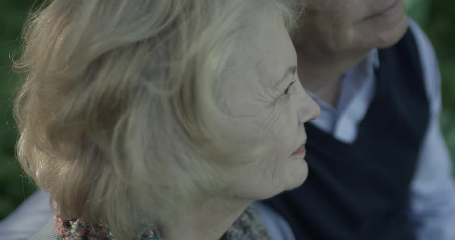 A man and a woman sit at a park bench they are both old and seem to be happy being with one another looking out at the park