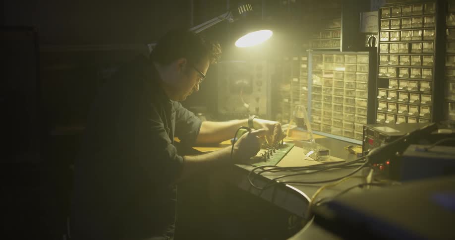A man solders something as he sits at a workbench with a single light