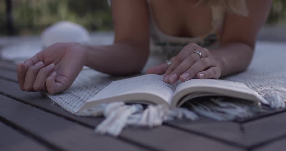The hands and arms of a woman laying on her stomach on a blanket and reading a book