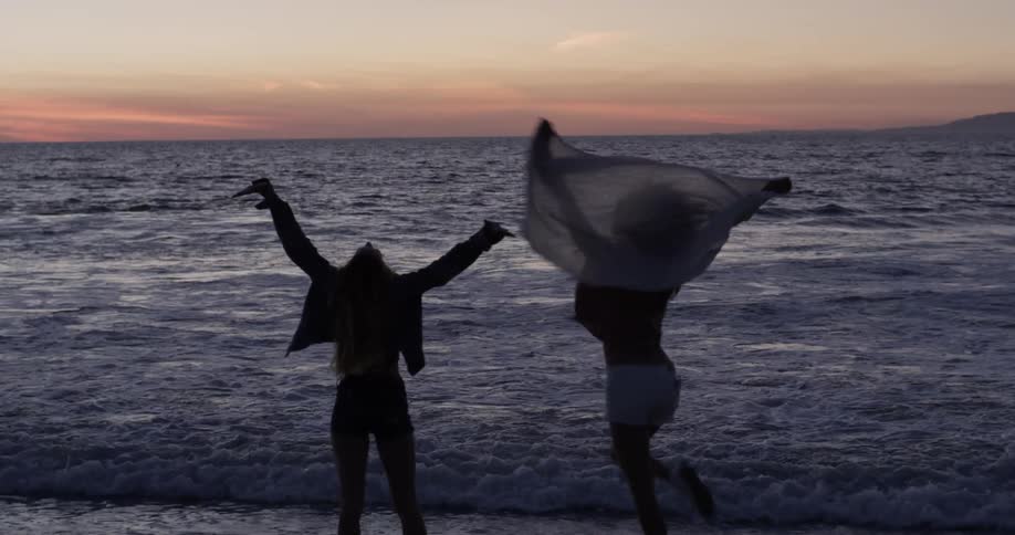 Two women run ankle deep into the ocean during a sunset