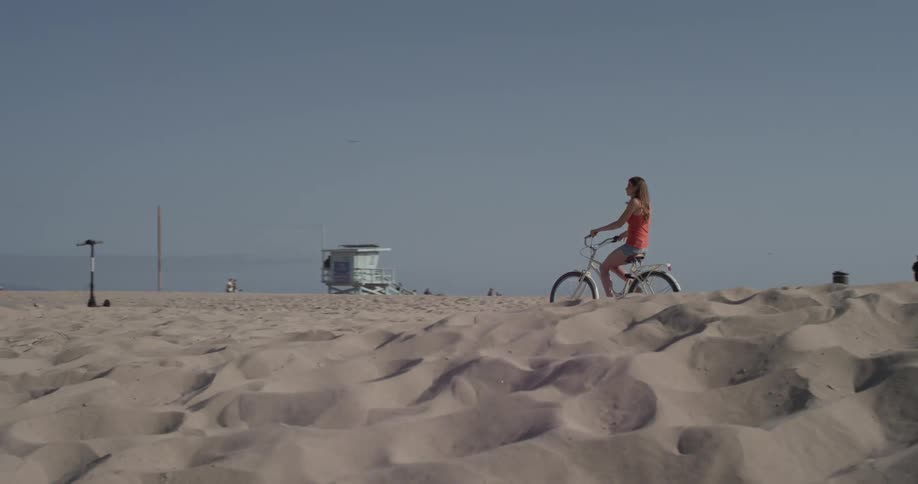 A woman peddles her bike along a path cutting through the sand at the beach
