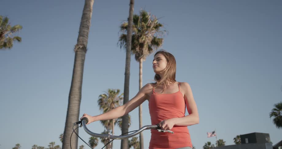 A woman walks her bike under a bright blue sky near palm trees