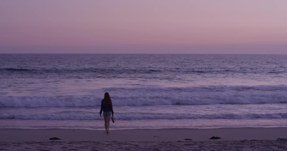 A woman holds her shoes and walks toward the ocean edge