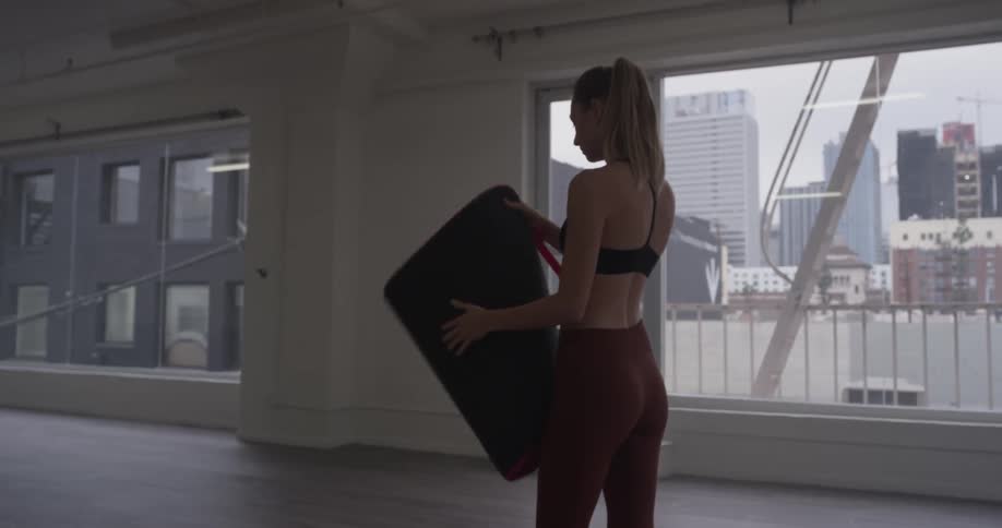 A woman unrolls a yoga mat in an empty studio with many windows