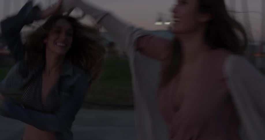 Two women dance together on a sidewalk after sunset with shaking booty shorts near the beach with palm trees around