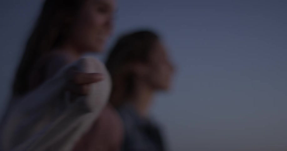 Two women at the beach are standing in the wind looking out at the distance and are very excited after the sun has set