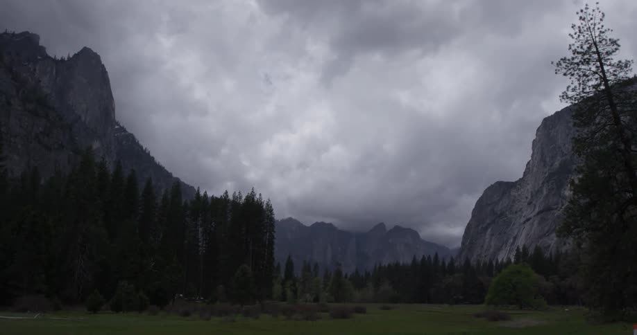 The clouds roll by on a dimly lit day in the mountains full of trees and grassy fields