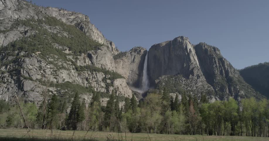 Water falls off a water fall from the top of a mountain off in the distance and trees sway in the wind a bit closer