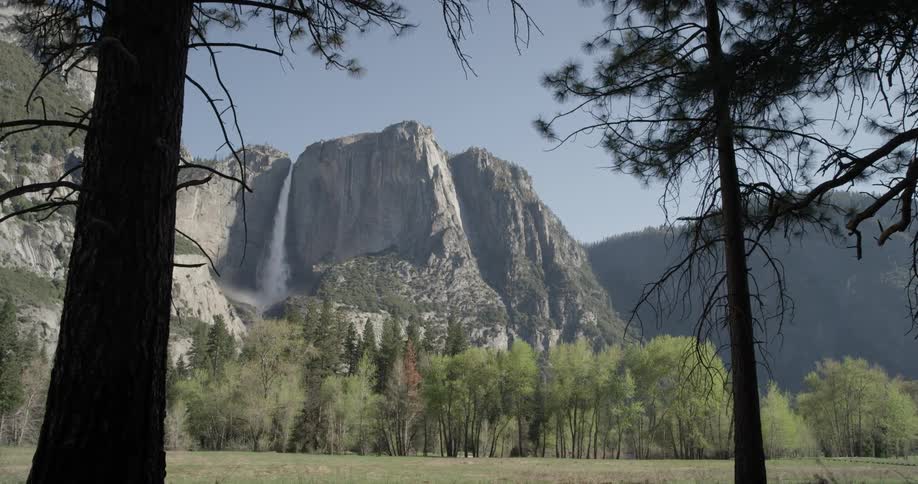 Water falls off of a mountain in the background while trees populate the grassy fields beneath on a sunny day